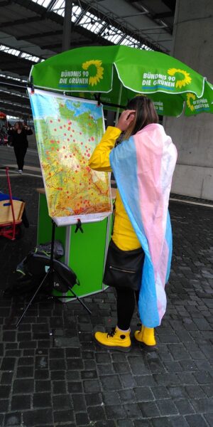 Mensch mit umgehängter Pride-Flagge steht vor einer Deutschlandkarte, auf der rote Punkte aufgeklebt sind