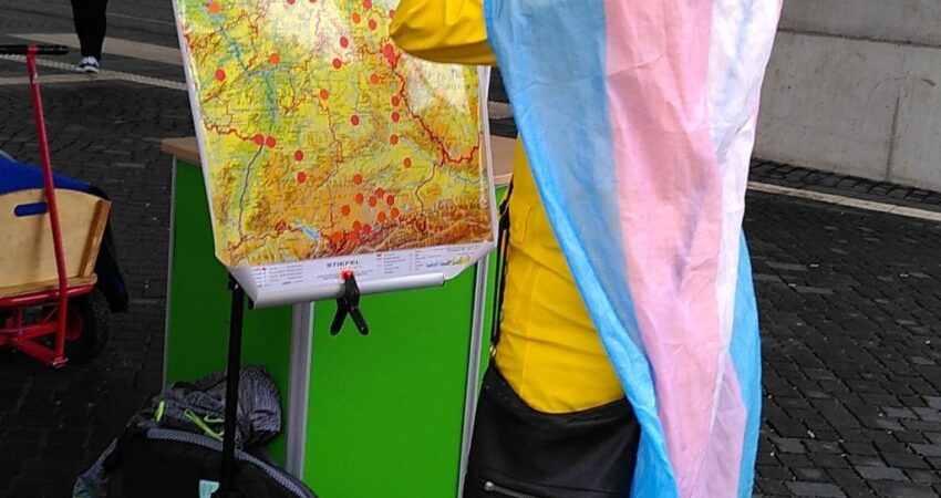 Mensch mit umgehängter Pride-Flagge steht vor einer Deutschlandkarte, auf der rote Punkte aufgeklebt sind